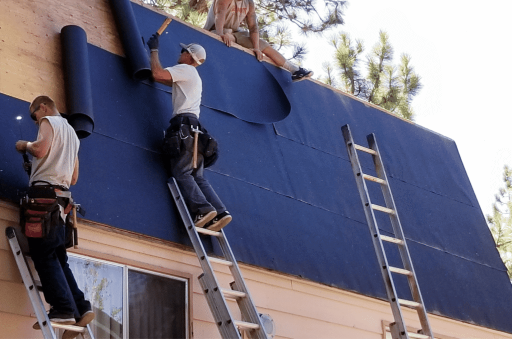 Roofers install black roof underlayment on a steep residential roof using ladders and safety equipment outdoors